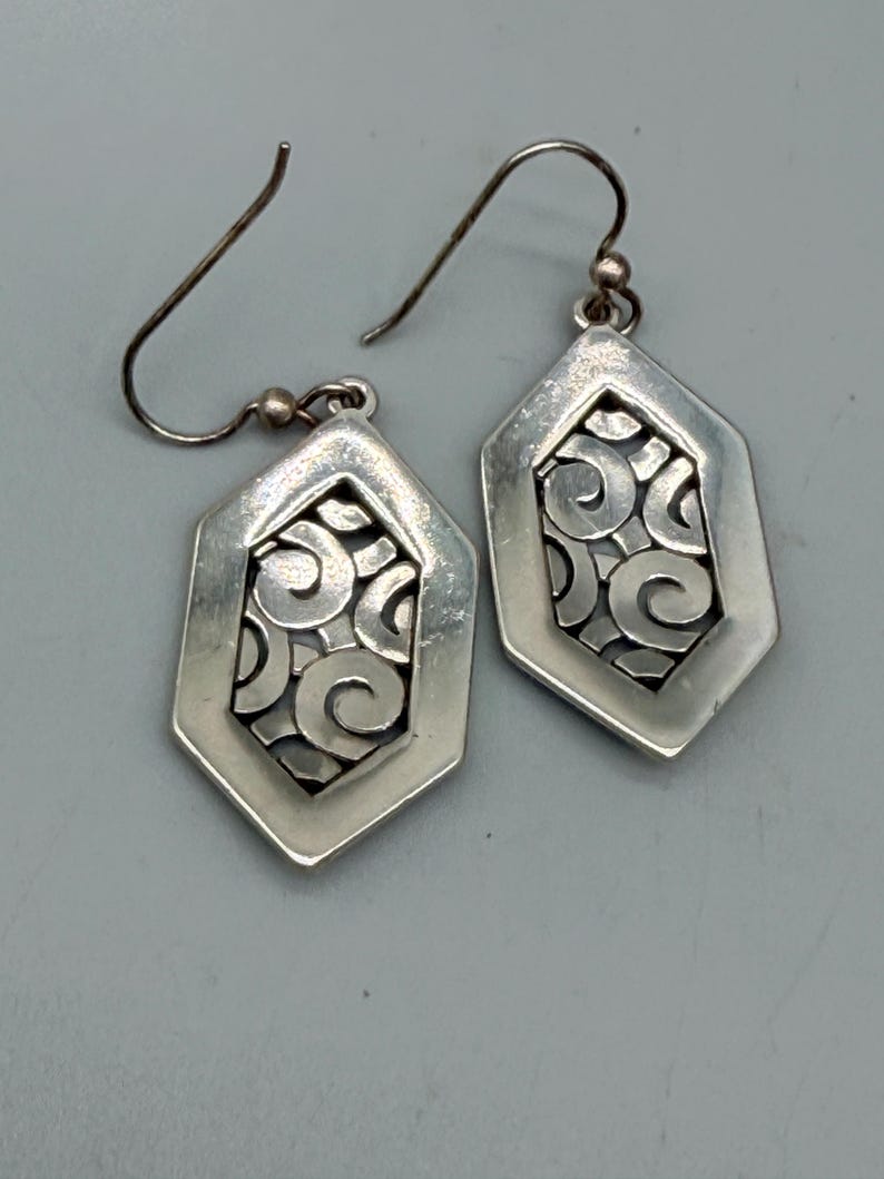 Puede incluir: Un par de pendientes plateados. Cada pendiente presenta una forma geom&eacute;trica hexagonal con un dise&ntilde;o calado decorativo de patrones arremolinados. Los pendientes tienen un cierre de gancho. El fondo es gris neutro.