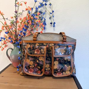 May include: A floral-patterned handbag with two front pockets and brown leather trim. The bag features a gray top and brown leather handles. A vase of colorful flowers sits to the left of the bag on a wooden surface.