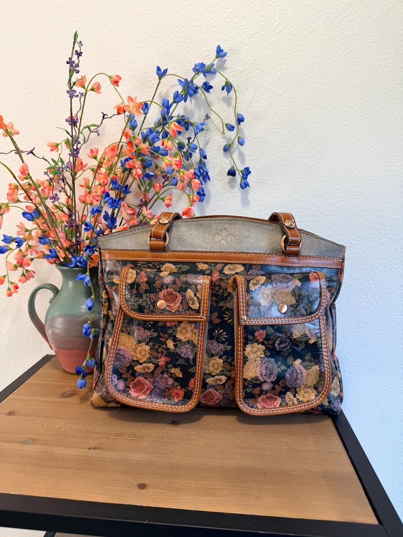 May include: A floral-patterned handbag with two front pockets and brown leather trim. The bag features a gray top and brown leather handles. A vase of colorful flowers sits to the left of the bag on a wooden surface.