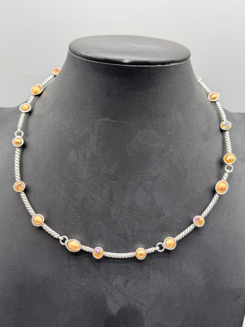 May include: A silver-toned necklace featuring a twisted cable chain design. The chain is accented with evenly spaced, round, amber-colored cabochon stones and smaller, iridescent, multi-colored faceted crystals set in circular silver-toned metal bezels.