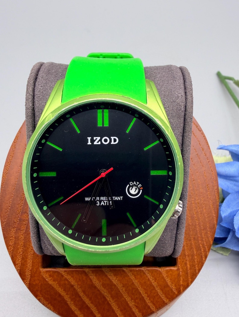 May include: An Izod wristwatch with a bright lime green silicone strap and matching bezel. The black dial features lime green hour markers, a red second hand, and a date window at the four o'clock position. The text reads IZOD and WATER RESISTANT 3 ATM.