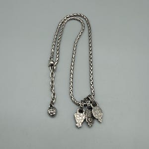 May include: A silver-toned necklace featuring a braided chain with an adjustable link section ending in a small heart-shaped charm. Three distinct, textured silver-toned pendants hang from the chain, each decorated with intricate heart patterns.