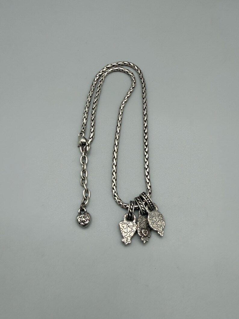 May include: A silver-toned necklace featuring a braided chain with an adjustable link section ending in a small heart-shaped charm. Three distinct, textured silver-toned pendants hang from the chain, each decorated with intricate heart patterns.