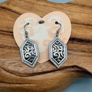 Puede incluir: Un par de pendientes plateados con un dise&ntilde;o geom&eacute;trico. Cada pendiente presenta un marco hexagonal incrustado con peque&ntilde;os cristales transparentes. El centro tiene un patr&oacute;n calado con detalles en espiral. Los pendientes cuelgan de un gancho simple.