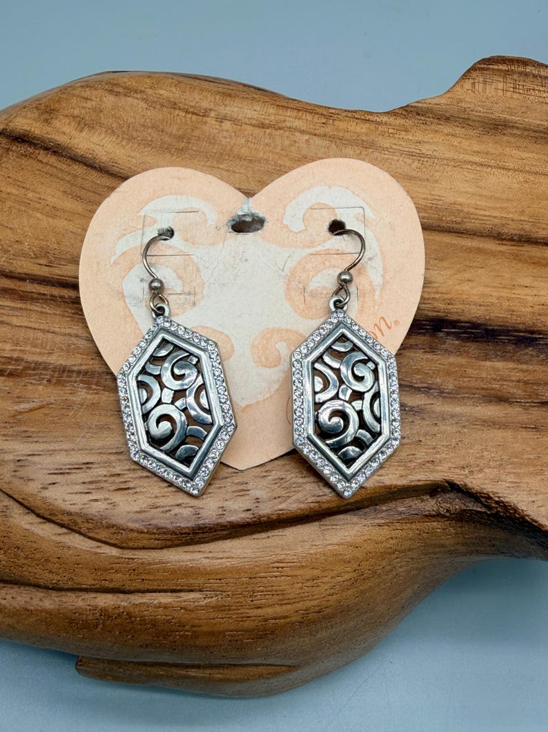 Puede incluir: Un par de pendientes plateados con un dise&ntilde;o geom&eacute;trico. Cada pendiente presenta un marco hexagonal incrustado con peque&ntilde;os cristales transparentes. El centro tiene un patr&oacute;n calado con detalles en espiral. Los pendientes cuelgan de un gancho simple.