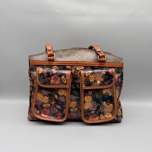 May include: A floral print tote bag with two front pockets. The bag features a dark background with a pattern of roses in shades of pink, yellow, and purple. The top and handles are brown leather, and the bag has a structured shape.