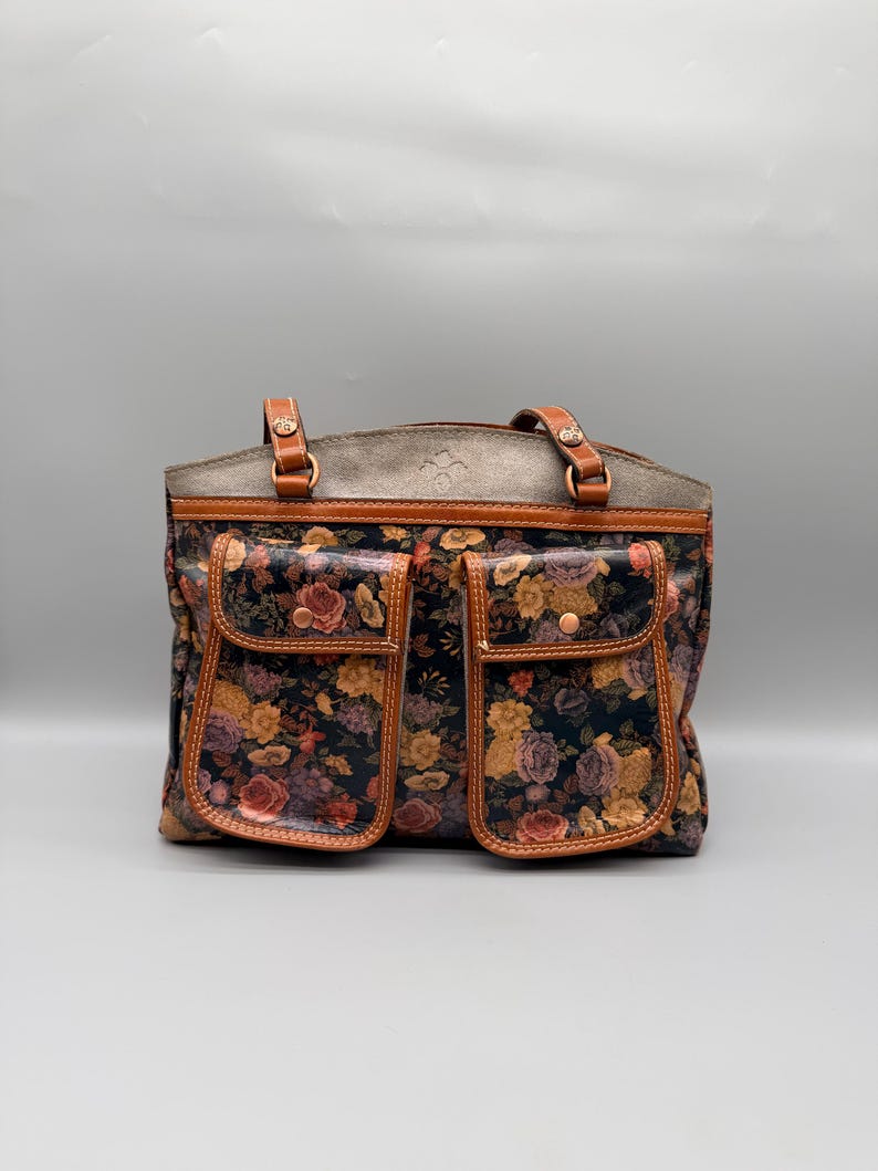 May include: A floral print tote bag with two front pockets. The bag features a dark background with a pattern of roses in shades of pink, yellow, and purple. The top and handles are brown leather, and the bag has a structured shape.
