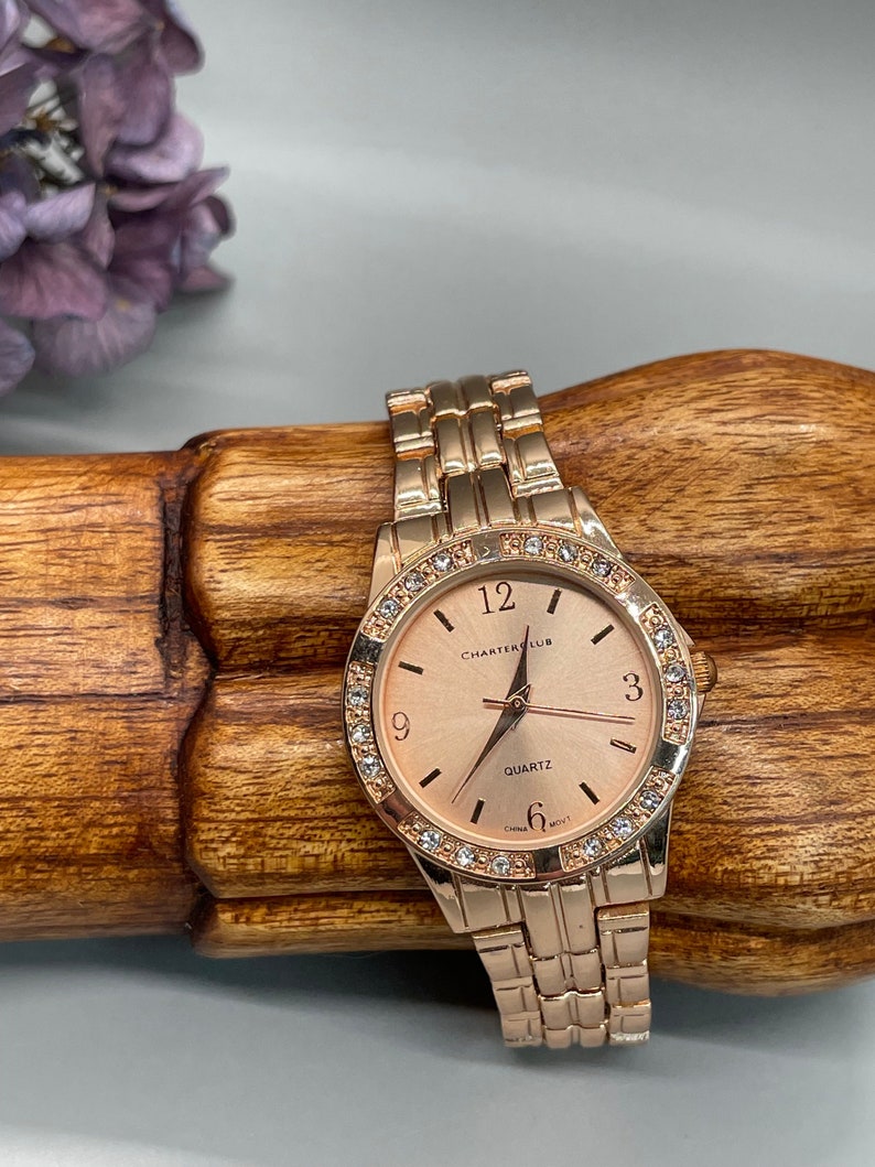 May include: A rose gold wristwatch with a rose gold band and a white face. The face has a rose gold trim and is adorned with small clear crystals. The watch face has black hands and black numbers. The watch face also has the words 'Charter Club' and 'Quartz' printed on it. The watch face also has the words 'China Mov't' printed on it.