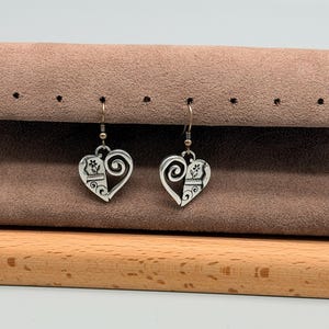 May include: A pair of silver-coloured heart-shaped earrings. Each earring features a decorative design with a paw print and floral elements. The earrings hang from silver-coloured fish hook ear wires. The earrings are displayed on a brown surface.