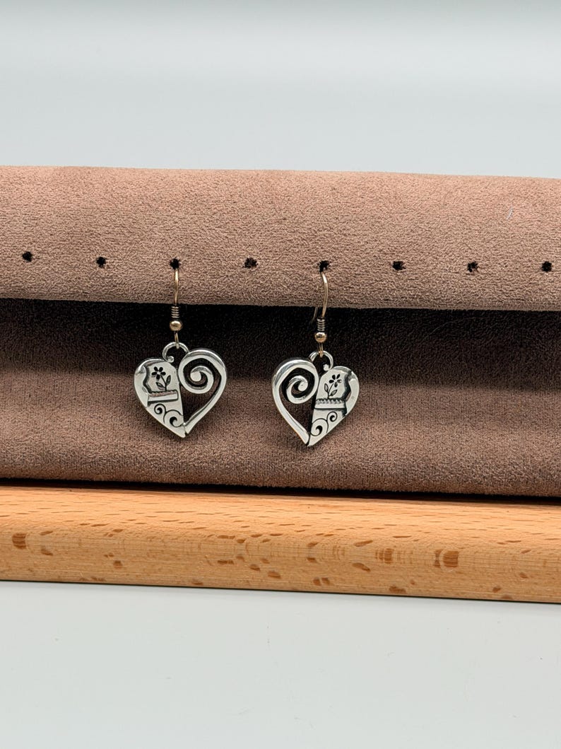 May include: A pair of silver-coloured heart-shaped earrings. Each earring features a decorative design with a paw print and floral elements. The earrings hang from silver-coloured fish hook ear wires. The earrings are displayed on a brown surface.