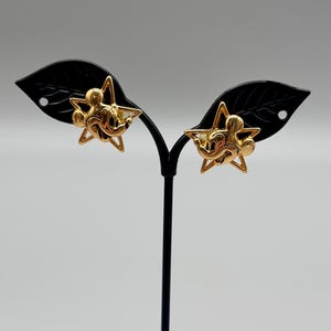 May include: A pair of gold-colored Mickey Mouse star-shaped earrings. The earrings feature a gold outline of a star with Mickey Mouse's face in the center. The earrings are displayed on a black earring stand against a light gray background.