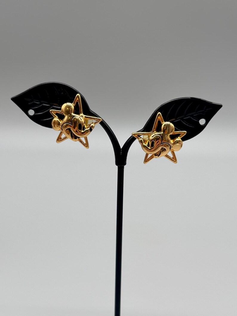 May include: A pair of gold-colored Mickey Mouse star-shaped earrings. The earrings feature a gold outline of a star with Mickey Mouse's face in the center. The earrings are displayed on a black earring stand against a light gray background.