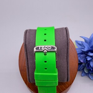 May include: A bright neon green silicone watch strap featuring a silver-toned metal buckle with the brand name IZOD engraved on the front. The strap includes a matching green silicone keeper loop and multiple adjustment holes for a secure fit.