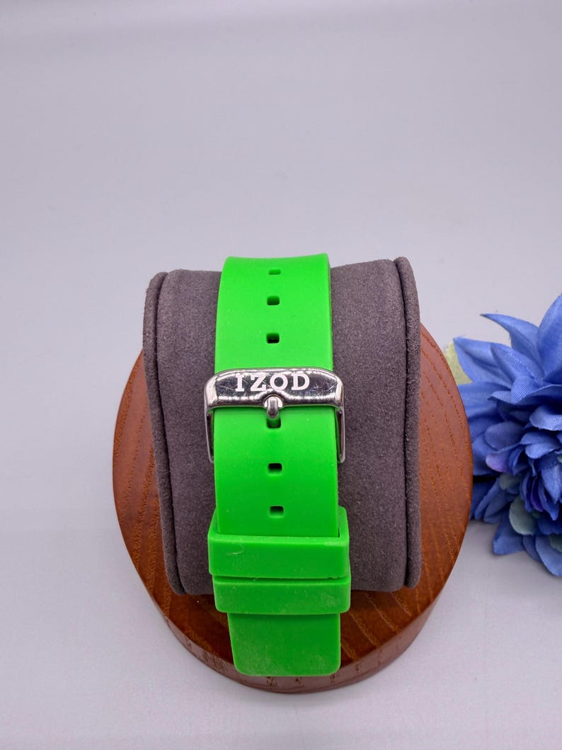 May include: A bright neon green silicone watch strap featuring a silver-toned metal buckle with the brand name IZOD engraved on the front. The strap includes a matching green silicone keeper loop and multiple adjustment holes for a secure fit.
