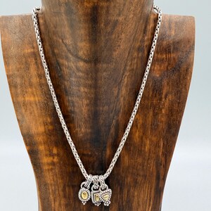 May include: A silver chain necklace featuring three small, dangling charms. Each charm has a textured silver border and a central gold-toned inlay. The charms are shaped as an oval, a square, and a triangle, all suspended from a braided silver chain.