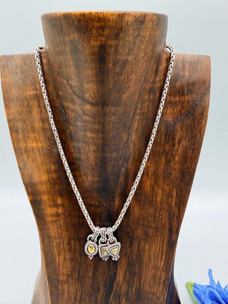 May include: A silver chain necklace featuring three small, dangling charms. Each charm has a textured silver border and a central gold-toned inlay. The charms are shaped as an oval, a square, and a triangle, all suspended from a braided silver chain.