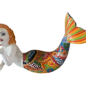 May include: A colorful ceramic mermaid figurine with a white body, red hair, and a multi-colored tail. The tail features a variety of geometric patterns and bright colors, including orange, yellow, green, blue, and red.