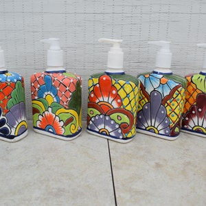 May include: Five colorful ceramic soap dispensers with floral designs. Each dispenser has a white pump and a square base. The dispensers are arranged in a row on a light gray surface.