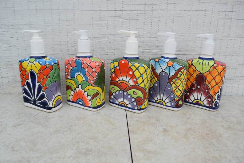 May include: Five colorful ceramic soap dispensers with floral designs. Each dispenser has a white pump and a square base. The dispensers are arranged in a row on a light gray surface.
