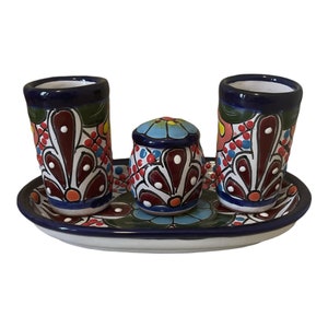 May include: A set of three ceramic shot glasses and a small jar with a blue and white floral design. The set is on a white oval tray with a blue rim and a floral design.