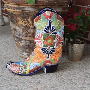 May include: A colorful ceramic cowboy boot planter with a floral design in shades of blue, green, orange, red, and white. The boot is decorated with a geometric pattern and has a blue base.
