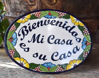 May include: Oval-shaped ceramic sign with a blue and white background. The sign has a colorful floral border and the text "Bienvenidos Mi Casa es su Casa" in blue cursive lettering.
