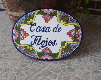 May include: A white oval ceramic sign with blue, yellow, and green floral designs. The sign reads "Casa de Flojos" in cursive.