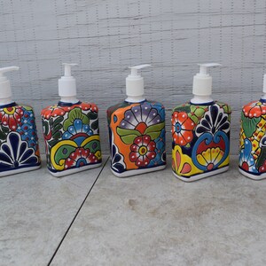 May include: Five colorful ceramic soap dispensers with floral patterns. Each dispenser has a white pump and a square base. The dispensers are arranged in a row on a gray tiled surface.