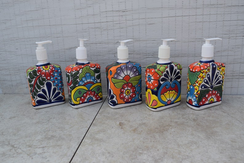 May include: Five colorful ceramic soap dispensers with floral patterns. Each dispenser has a white pump and a square base. The dispensers are arranged in a row on a gray tiled surface.
