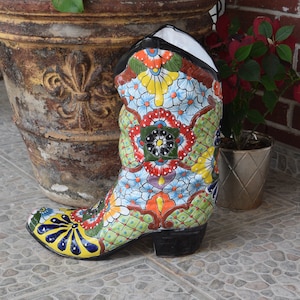 May include: A colorful ceramic boot planter with a floral design. The boot is painted with a variety of colors, including blue, green, yellow, red, and orange. The boot is sitting on a tiled floor.