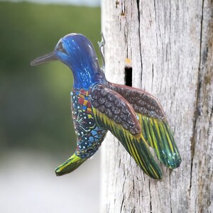 May include: A blue, green, and brown hummingbird wind chime hangs from a wooden post. The hummingbird is made of metal and has intricate details.
