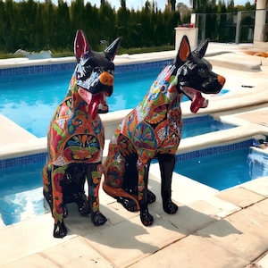 May include: Two decorative dog statues with black bodies and colorful floral patterns. The statues have their tongues out and are positioned near a swimming pool. The dogs have red tongues, orange noses, and red and orange ears.
