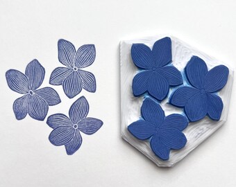 May include: A blue floral stamp with three stamped impressions of the design. The stamp itself features four blue flower shapes. The stamped impressions are also blue flowers, each with detailed line work, on a white background.