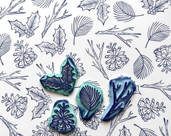 May include: Four blue and green rubber stamps with designs of holly leaves, pine cones, and branches. The stamps are on a white background with a repeating pattern of blue line drawings of holly leaves, pine cones, and branches.