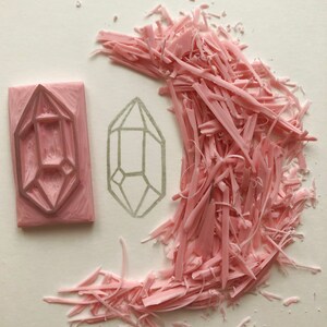 May include: A pink rubber stamp with a geometric design of a crystal. The stamp has been used and there are pink rubber shavings around it.