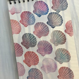 Op de afbeelding: Een notitieboek met een patroon van kleurrijke schelpen die op de pagina's zijn gedrukt. De schelpen zijn in tinten roze, paars, blauw en groen.