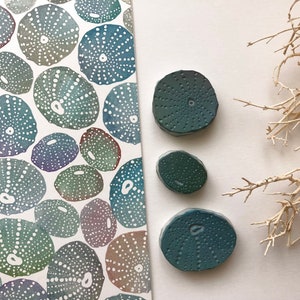 May include: Three teal blue sea urchin shaped ceramic coasters with a textured surface. The coasters are arranged in a triangle shape on a white surface.