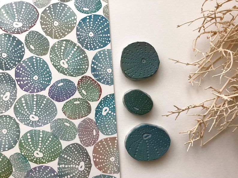 May include: Three teal blue sea urchin shaped ceramic coasters with a textured surface. The coasters are arranged in a triangle shape on a white surface.