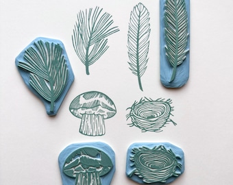 May include: Set of eight rubber stamps featuring nature-themed designs. The stamps are mounted on blue blocks and feature images of a mushroom, a nest, and various leaves and feathers.