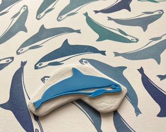 May include: A blue rubber stamp of a dolphin with a white background. The stamp is surrounded by a pattern of blue dolphins printed on a white surface.