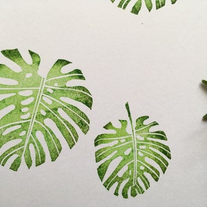 Puede incluir: Un fondo blanco con cuatro estampados de hojas verdes en acuarela. Las hojas son una versión estilizada de una hoja de monstera.
