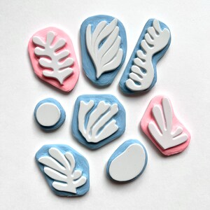 K&ouml;nnte beinhalten: Ein Satz von acht Stempeln aus Gummi mit abstrakten Blatt- und Blumenmotiven. Die Stempel sind in verschiedenen Formen und Gr&ouml;&szlig;en, einige sind rosa und andere blau.