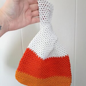 May include: A crochet wristlet bag featuring a color-blocked design with a white top section, a vibrant orange middle band, and a golden-yellow base. The bag has a single integrated handle loop and a triangular shape resembling a candy corn piece.