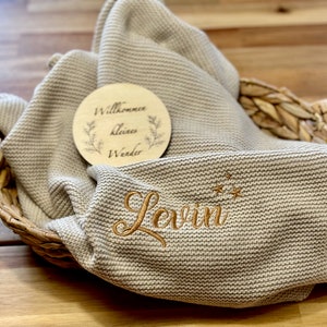 Könnte beinhalten: Eine beige gestrickte Babydecke mit dem Namen "Levin" in goldener Schrift gestickt und drei goldenen Sternen über dem Namen. Ein Holzschild mit dem Text "Willkommen kleines Wunder" liegt auf der Decke.