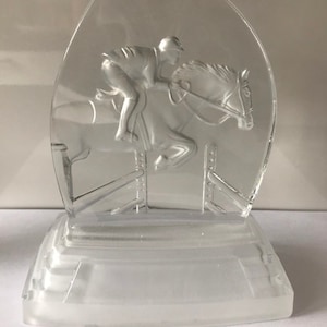 May include: A clear glass sculpture of a horse jumping over a hurdle with a rider on its back. The sculpture is on a clear glass base.