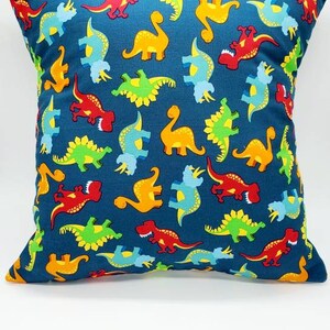 May include: A blue pillow with a pattern of colorful dinosaurs. The dinosaurs are in various poses, including standing, walking, and running. The pillow is made of a soft, comfortable fabric.