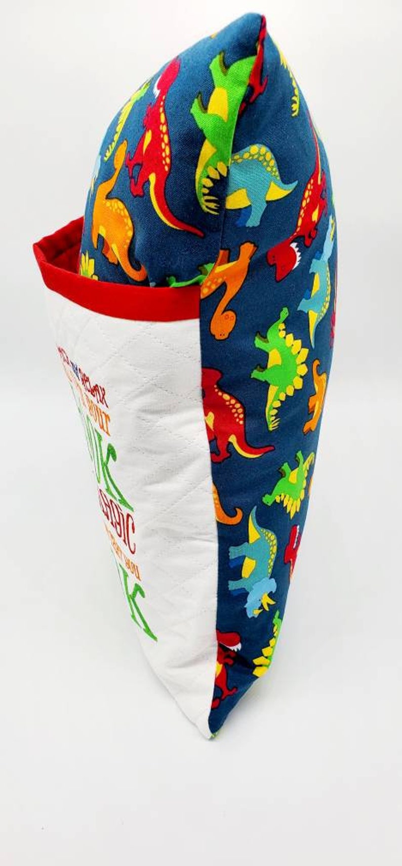 May include: A white pillow with red trim and a blue fabric back featuring a repeating pattern of colorful dinosaurs. The front of the pillow has the text "You are stronger than you think, about it, you are magic, just let you, be you".
