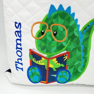 May include: A white quilted pillow with a green dinosaur wearing glasses and reading a book. The dinosaur has blue spikes on its back and blue paw prints on its feet. The text "Thomas" is embroidered on the pillow.