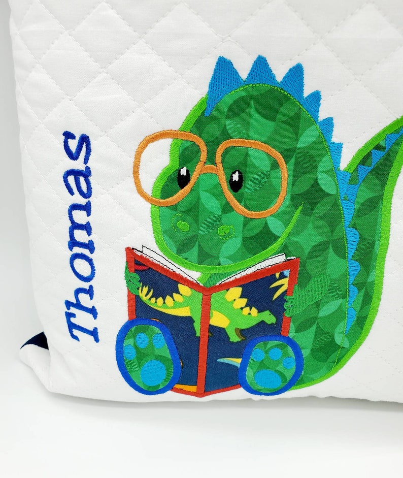 May include: A white quilted pillow with a green dinosaur wearing glasses and reading a book. The dinosaur has blue spikes on its back and blue paw prints on its feet. The text "Thomas" is embroidered on the pillow.