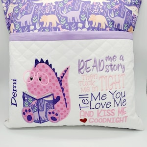 May include: A purple and white pillow with a dinosaur reading a book. The pillow has a pocket on the front and the text "READ me a story Then TUCK Me in TIGHT Tell Me You Love Me AND KISS ME GOODNIGHT". The name "Demi" is embroidered on the side of the pillow.
